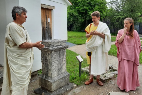 Im Tempelbezirk: Zwei Männer und eine Frau in römischem Gewand stehen vor einem Weihestein und unterhalten sich.