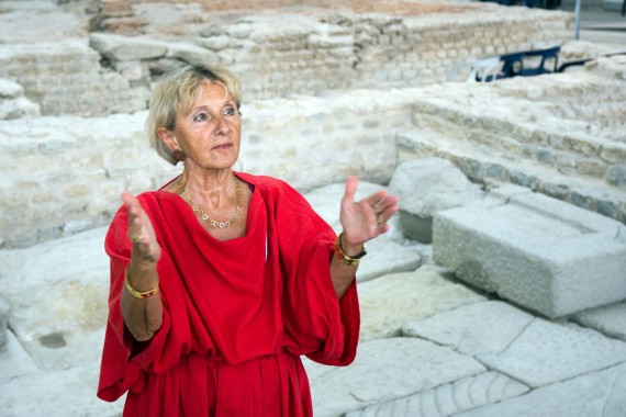 Frau in roter Tunica gestikuliert vor historischem Gemäuer der römischen Badeanstalt in den Kleinen Thermen im APC