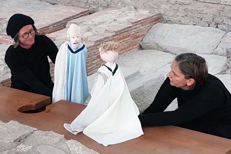 Zwei schwarz gekleidete Frauen mit Handpuppen in r&ouml;mischem Gewand auf der Latrine