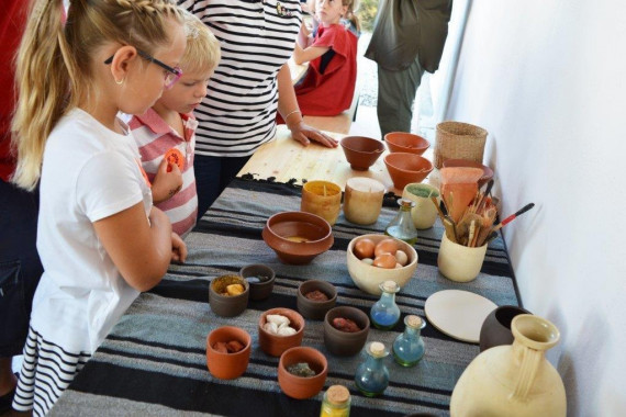 Zwei Kinder an Tisch mit Tongefäßen und Fläschchen. Sie sind mit unterschiedlichen Inhalten (Blüten, Pulver, Eier) gefüllt