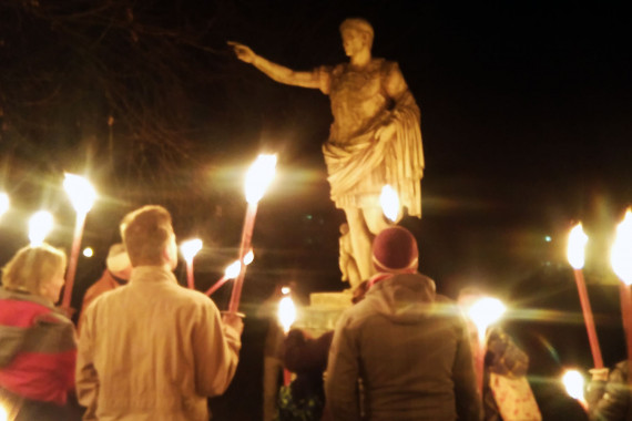 Es ist dunkel. Menschengruppe mit leuchtenden Fackeln stehen vor Augustus-Denkmal.