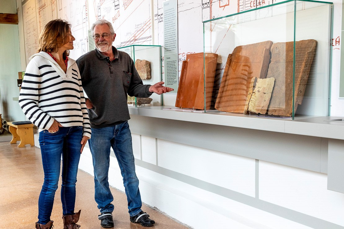 Exhibition &quot;For God's sake&quot;: Man and woman talking at a display case with roof tiles from the Roman city of Cambodunum