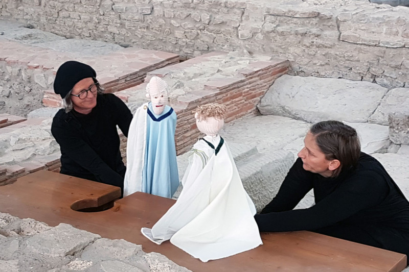 Zwei schwarz gekleidete Frauen mit Handpuppen in römischem Gewand auf der Latrine