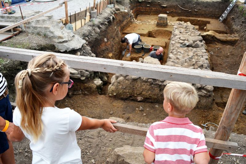 Zwei Kinder blicken in eine Ausgrabung, es sind Arch&auml;ologen bei der Arbeit sichtbar
