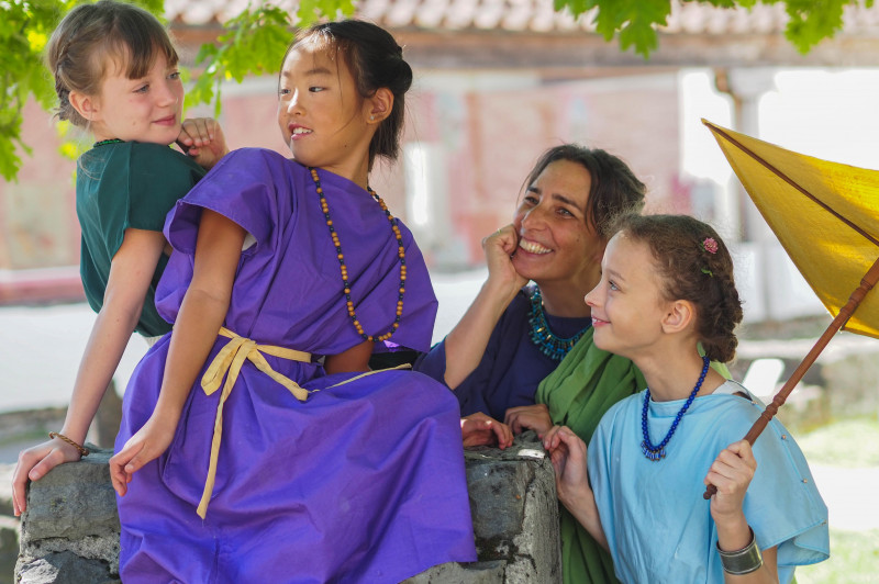 Drei M&auml;dchen und eine Frau in r&ouml;mischer Kleidung stehen im Tempelbezirk des APC vertraut beieinander 