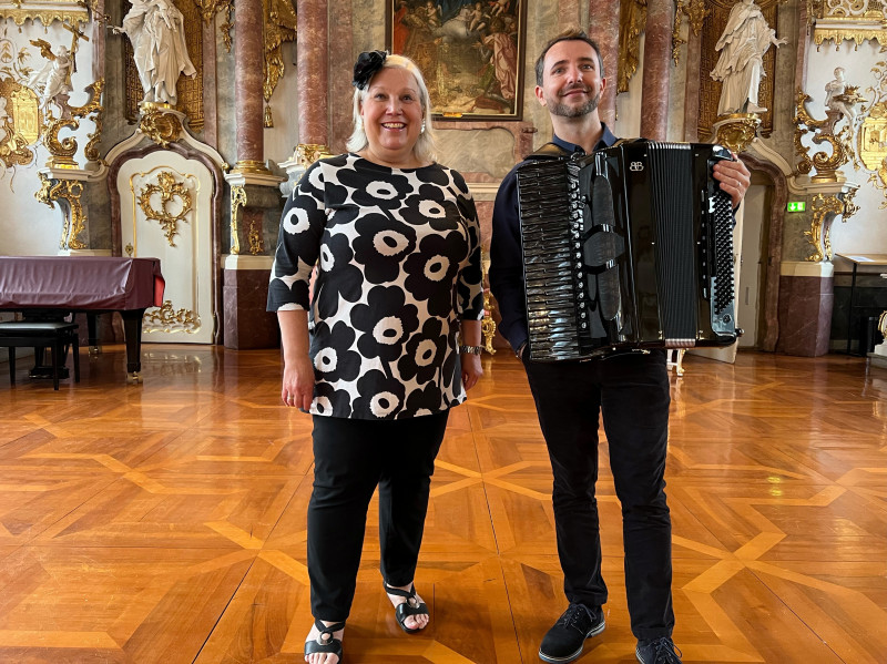 Das Duo, ein Mann und eine Frau, posieren im barocken Festsaal mit einem Akkordeon