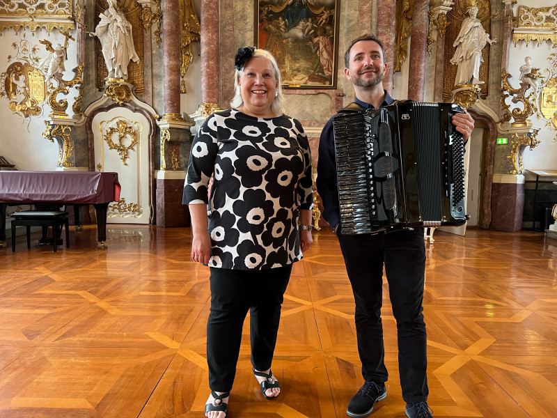 Das Duo, ein Mann und eine Frau, posieren im barocken Festsaal mit einem Akkordeon